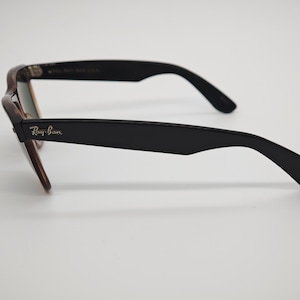 May include: A pair of Ray-Ban sunglasses with a tortoiseshell frame and black arms. The Ray-Ban logo is visible on the left arm. The lenses have a slight green tint.