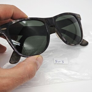 Puede incluir: Gafas de sol con montura oscura y lentes verdes. Las monturas tienen un aspecto moteado y texturizado. Las gafas de sol est&aacute;n sujetas por una mano y se ve una peque&ntilde;a etiqueta blanca con el n&uacute;mero "503".