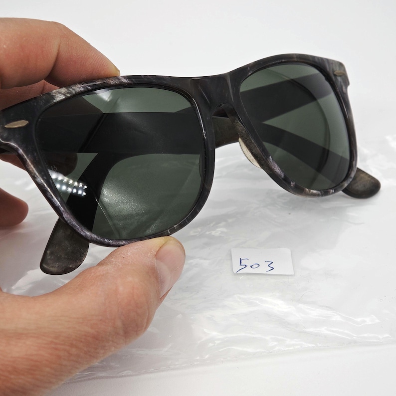 Puede incluir: Gafas de sol con montura oscura y lentes verdes. Las monturas tienen un aspecto moteado y texturizado. Las gafas de sol est&aacute;n sujetas por una mano y se ve una peque&ntilde;a etiqueta blanca con el n&uacute;mero "503".