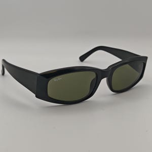 Może przedstawiać: Czarne okulary przeciwsłoneczne z prostokątnymi oprawkami i zielonymi soczewkami. Logo Ray-Ban jest widoczne na prawej soczewce. Okulary przeciwsłoneczne mają elegancki, nowoczesny design, odpowiedni do codziennego użytku.