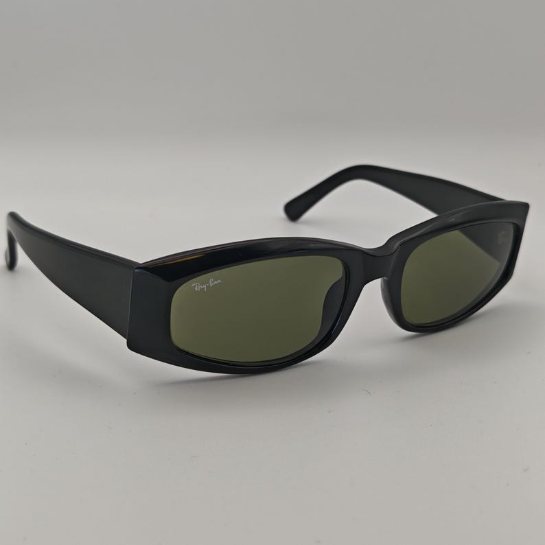 Może przedstawiać: Czarne okulary przeciwsłoneczne z prostokątnymi oprawkami i zielonymi soczewkami. Logo Ray-Ban jest widoczne na prawej soczewce. Okulary przeciwsłoneczne mają elegancki, nowoczesny design, odpowiedni do codziennego użytku.