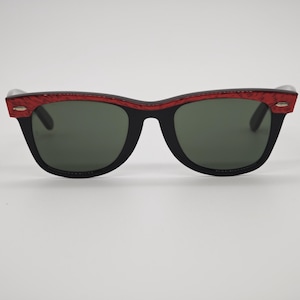 May include: Black sunglasses with a red patterned top frame and dark green lenses. The sunglasses have a classic, rectangular shape. The frame is black with a red textured pattern on the top. The lenses are a dark green colour.