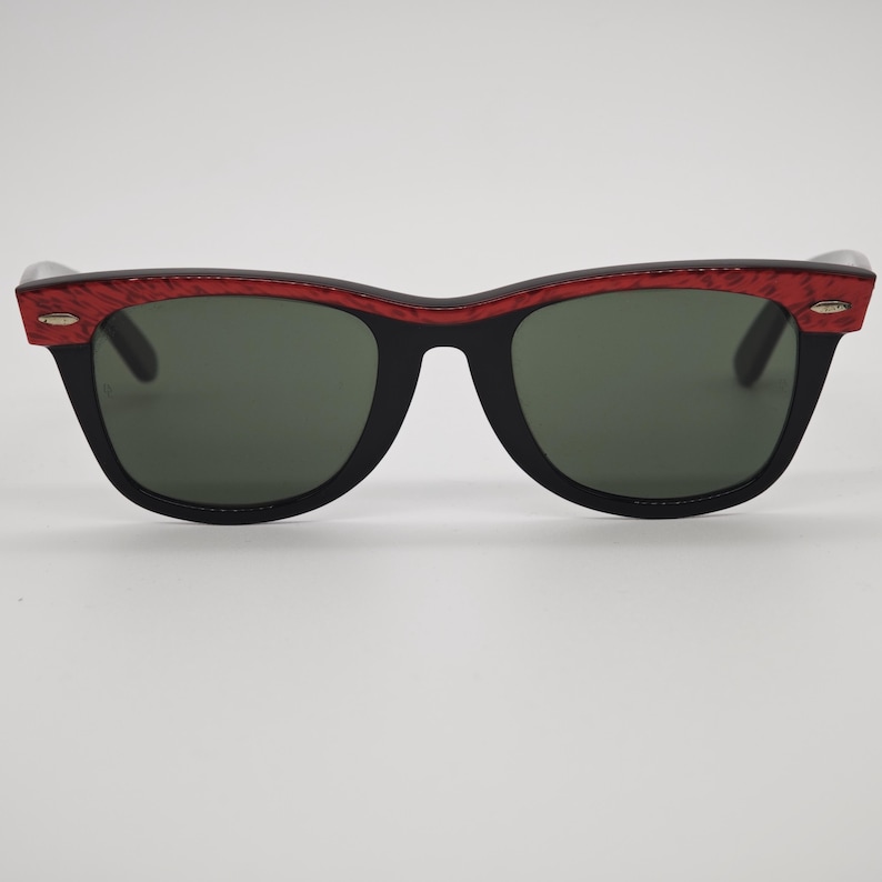 May include: Black sunglasses with a red patterned top frame and dark green lenses. The sunglasses have a classic, rectangular shape. The frame is black with a red textured pattern on the top. The lenses are a dark green colour.