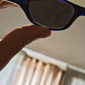 May include: Sunglasses with a dark lens and a dark blue frame. The sunglasses are held up by a hand. The background is blurred.