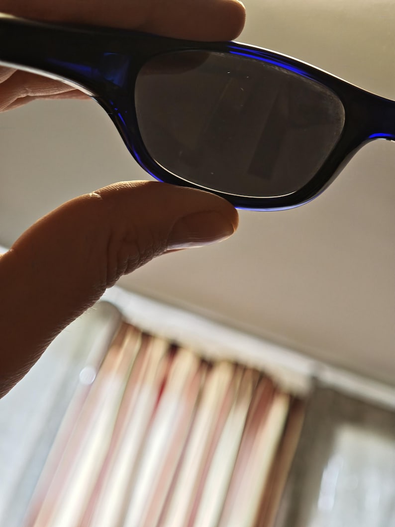 May include: Sunglasses with a dark lens and a dark blue frame. The sunglasses are held up by a hand. The background is blurred.