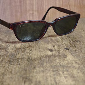 May include: Tortoiseshell-framed sunglasses with dark lenses rest on a wooden surface. The sunglasses have a classic design with a slightly curved frame and the Ray-Ban logo visible on the lens. The overall aesthetic is vintage and stylish.