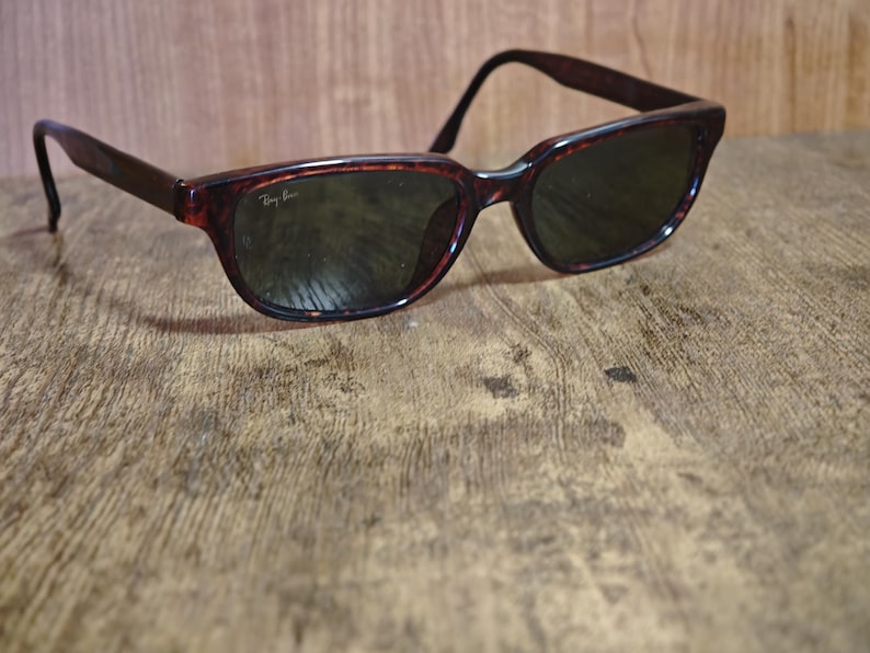 May include: Tortoiseshell-framed sunglasses with dark lenses rest on a wooden surface. The sunglasses have a classic design with a slightly curved frame and the Ray-Ban logo visible on the lens. The overall aesthetic is vintage and stylish.