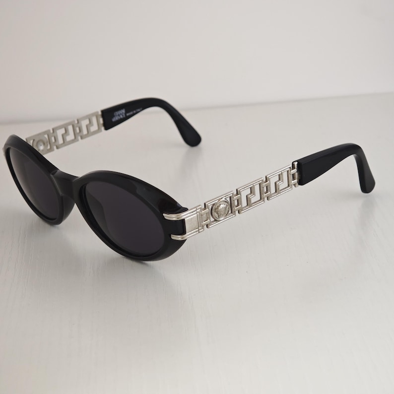 Puede incluir: Gafas de sol ovaladas negras con patillas de metal plateado con un patr&oacute;n geom&eacute;trico griego y un emblema circular de cabeza de Medusa. Las monturas de pl&aacute;stico negro tienen lentes oscuras y la patilla interior muestra Gianni Versace.