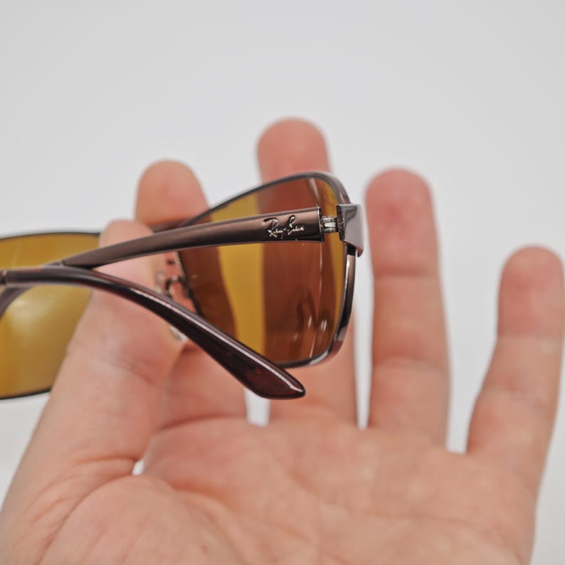 May include: Brown sunglasses with a Ray-Ban logo on the temple. The lenses are a warm brown color, and the frames are a darker brown. The sunglasses are held in a hand against a white background.