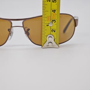 May include: Brown aviator sunglasses with amber-colored lenses. The sunglasses are being measured with a yellow tape measure. The frame is a dark brown color. The lenses are a light amber color.