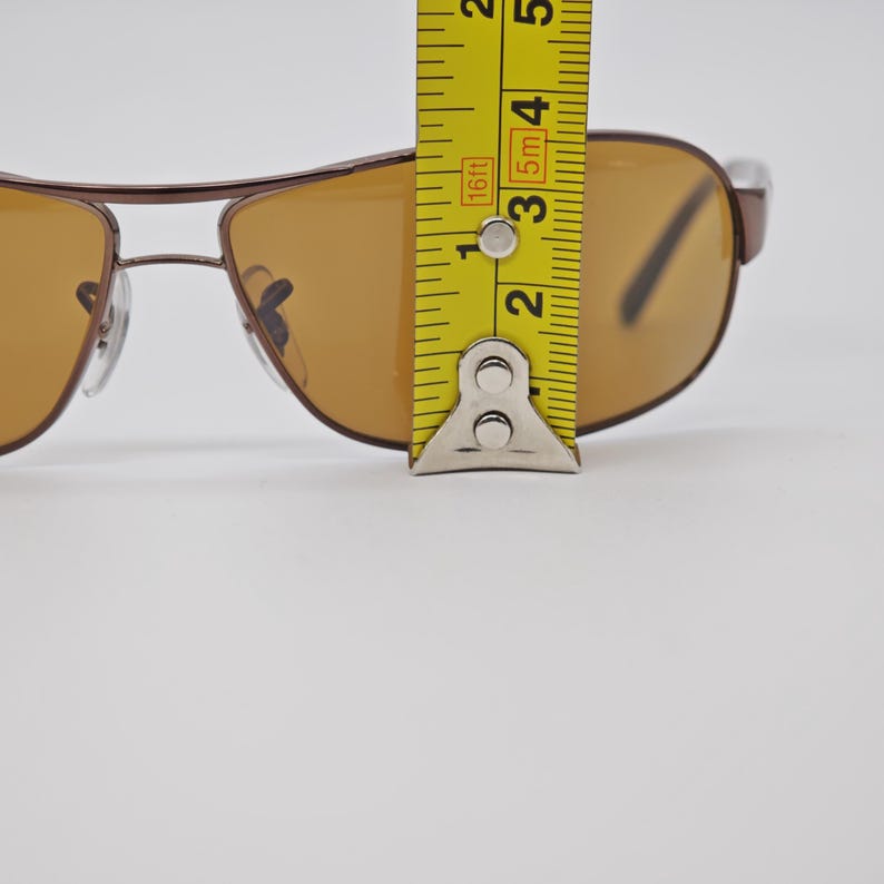 May include: Brown aviator sunglasses with amber-colored lenses. The sunglasses are being measured with a yellow tape measure. The frame is a dark brown color. The lenses are a light amber color.