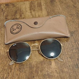 May include: Gold-rimmed sunglasses with dark lenses and a matching tan leather case. The sunglasses have a classic round shape and the case is closed with a button. The Ray-Ban logo is visible on the sunglasses and the case.