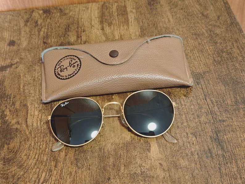 May include: Gold-rimmed sunglasses with dark lenses and a matching tan leather case. The sunglasses have a classic round shape and the case is closed with a button. The Ray-Ban logo is visible on the sunglasses and the case.