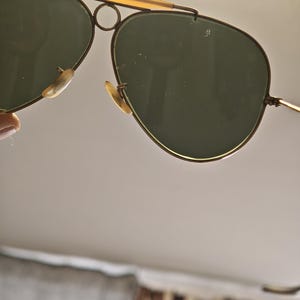 May include: Aviator-style sunglasses with a gold-tone metal frame and dark green lenses. The sunglasses have a double bridge and a curved brow bar. The lenses are teardrop-shaped, and the nose pads are a light beige color.