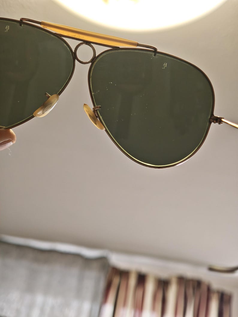 May include: Aviator-style sunglasses with a gold-tone metal frame and dark green lenses. The sunglasses have a double bridge and a curved brow bar. The lenses are teardrop-shaped, and the nose pads are a light beige color.
