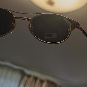 May include: Gold-framed sunglasses with dark lenses. The sunglasses have a double bridge and a classic, rounded shape. The lenses appear to be slightly reflective, and the frame has a subtle, textured finish. The overall design is vintage-inspired.