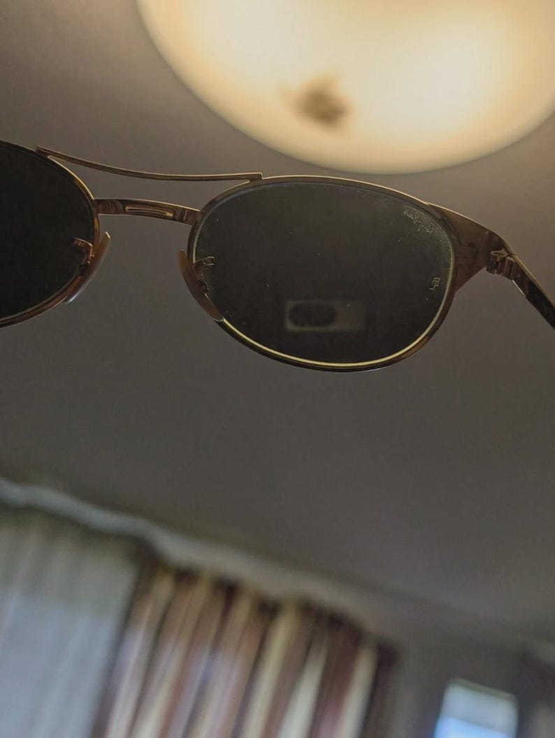 May include: Gold-framed sunglasses with dark lenses. The sunglasses have a double bridge and a classic, rounded shape. The lenses appear to be slightly reflective, and the frame has a subtle, textured finish. The overall design is vintage-inspired.