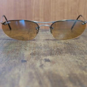 May include: Rimless sunglasses with a silver metal frame and brown tinted lenses. The Ray-Ban logo is visible on the upper left lens. The sunglasses are resting on a wooden surface.