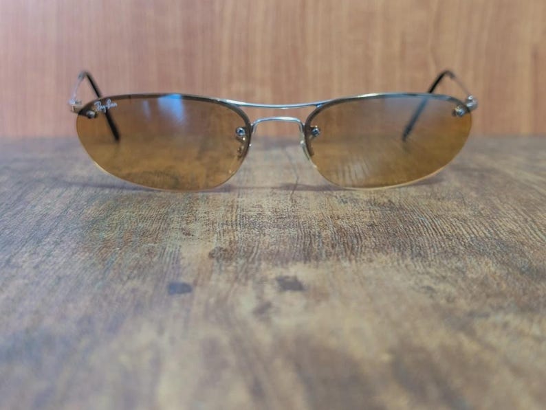 May include: Rimless sunglasses with a silver metal frame and brown tinted lenses. The Ray-Ban logo is visible on the upper left lens. The sunglasses are resting on a wooden surface.