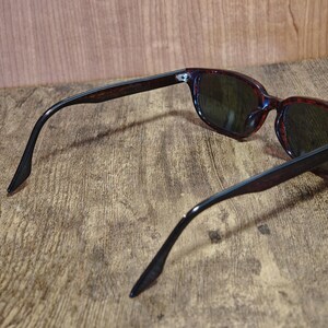 May include: Sunglasses with dark lenses and a tortoiseshell pattern frame. The arms are a dark brown color. The sunglasses are resting on a wooden surface.