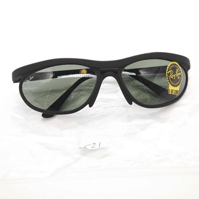 May include: Black Ray-Ban sunglasses with a curved frame and green-tinted lenses. The sunglasses have a small Ray-Ban logo on the left arm and a gold sticker on the right lens. The lenses are oval-shaped. The sunglasses are on a white surface.