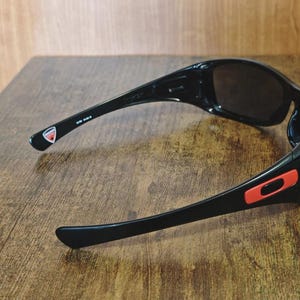 K&ouml;nnte beinhalten: Schwarze Sonnenbrille mit dunklen Gl&auml;sern. Die Sonnenbrille hat ein schlankes, sportliches Design mit einem roten Akzent an der Seite und einem Logo am B&uuml;gel. Die Sonnenbrille liegt auf einer Holzoberfl&auml;che.