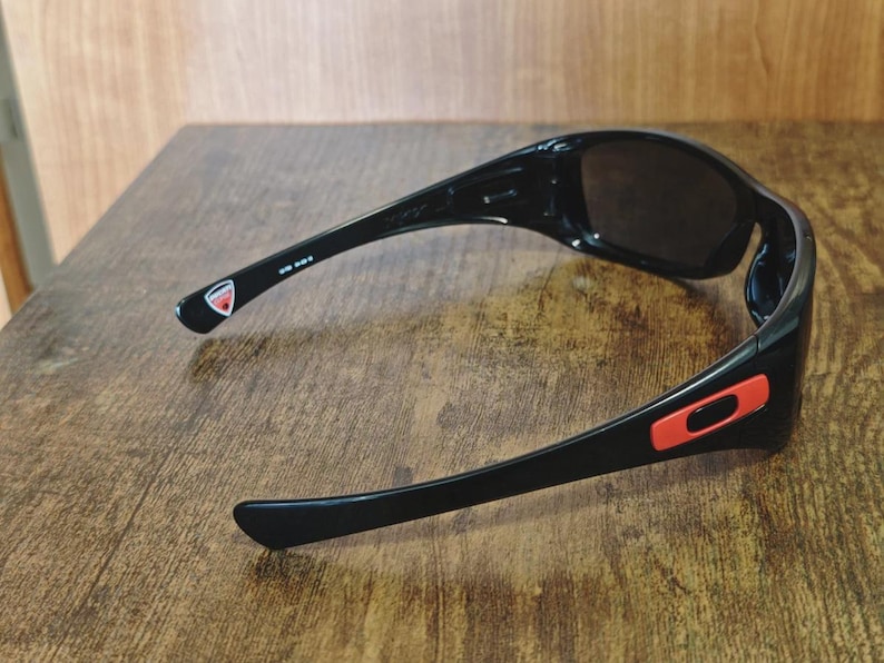 K&ouml;nnte beinhalten: Schwarze Sonnenbrille mit dunklen Gl&auml;sern. Die Sonnenbrille hat ein schlankes, sportliches Design mit einem roten Akzent an der Seite und einem Logo am B&uuml;gel. Die Sonnenbrille liegt auf einer Holzoberfl&auml;che.