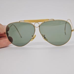 May include: Gold-framed aviator sunglasses with green lenses. The sunglasses feature a double bridge and a light-colored bar above the lenses. The lenses are teardrop-shaped, and the frame has a classic, vintage design.