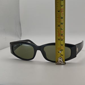 Może przedstawiać: Czarne okulary przeciwsłoneczne z zielonymi soczewkami. Okulary mają prostokątny kształt i małe logo po lewej stronie. Okulary mają około 13 cm długości.