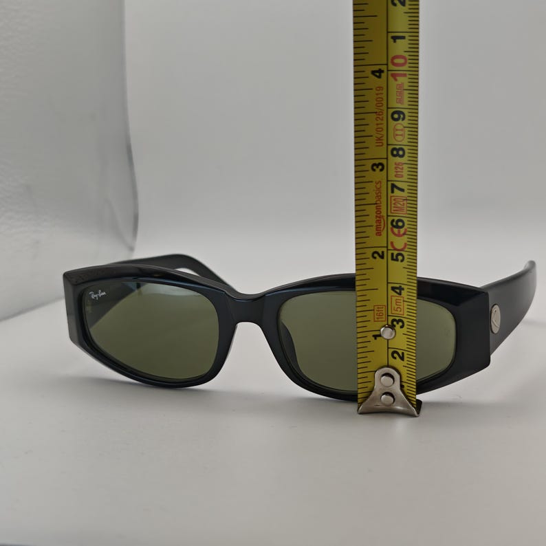 Może przedstawiać: Czarne okulary przeciwsłoneczne z zielonymi soczewkami. Okulary mają prostokątny kształt i małe logo po lewej stronie. Okulary mają około 13 cm długości.