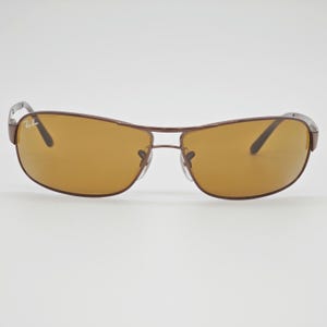 May include: Brown sunglasses with a rectangular frame and amber lenses. The sunglasses have a metal frame and the Ray-Ban logo is visible on the left lens. The sunglasses are designed for outdoor use and provide protection from the sun.