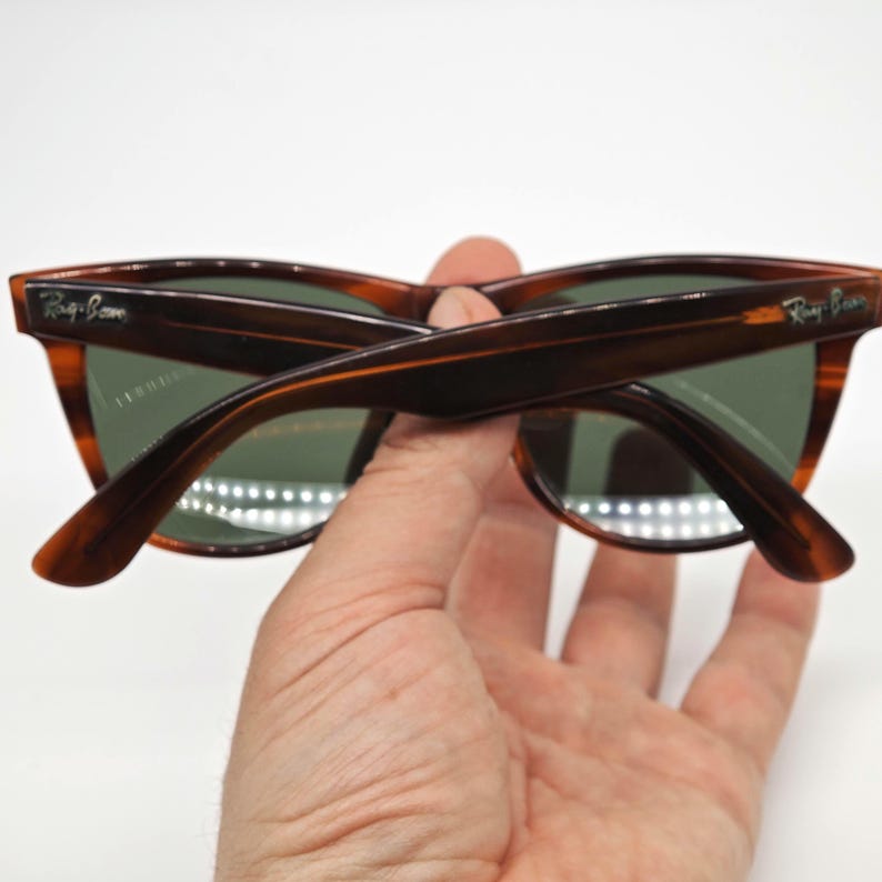 May include: Tortoiseshell-patterned sunglasses with green lenses. The frames have a classic design, and the Ray-Ban logo is visible on the temples. The sunglasses are held in a hand against a white background.