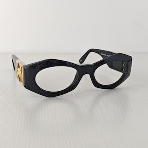 以下が含まれることがあります： テンプルにゴールドトーンの金属製アクセントをあしらった、黒い幾何学模様のメガネ。フレームは厚みのある光沢のある黒いプラスチック製で、独特の角張ったデザインとサイドのヒンジ部分に目立つゴールドのエンブレムが特徴です。