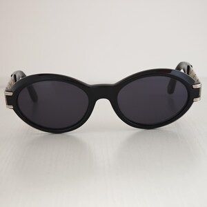 Puede incluir: Gafas de sol de forma ovalada con una montura negra gruesa y lentes oscuras. Las patillas presentan detalles met&aacute;licos en tono plateado en las bisagras, lo que proporciona un contraste distintivo frente a la estructura de pl&aacute;stico negro s&oacute;lido.