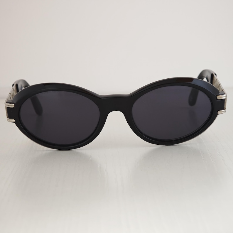 Puede incluir: Gafas de sol de forma ovalada con una montura negra gruesa y lentes oscuras. Las patillas presentan detalles met&aacute;licos en tono plateado en las bisagras, lo que proporciona un contraste distintivo frente a la estructura de pl&aacute;stico negro s&oacute;lido.