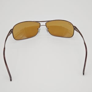 May include: Brown aviator sunglasses with amber-colored lenses. The sunglasses have a double bridge and a thin, dark brown metal frame. The temples are also dark brown. The Ray-Ban logo is visible on the right lens.