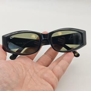 Może przedstawiać: Czarne okulary przeciwsłoneczne Ray-Ban Rituals z prostokątną oprawką i ż&oacute;łtymi szkłami. Okulary przeciwsłoneczne mają logo Ray-Ban po lewej stronie i napis "RAY-BAN RITUALS" na lewej soczewce. Zausszniki są czarne ze złotymi akcentami.