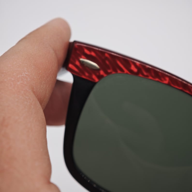 May include: Close-up of sunglasses with a black frame and green lenses. The top of the frame has a red, textured pattern. A small, gold-coloured accent is visible on the frame.