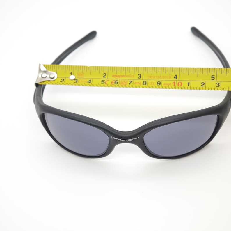 May include: Black sunglasses with gray lenses are measured with a yellow tape measure. The sunglasses have a curved frame and the brand name "Oakley" is visible on the frame. The sunglasses measure approximately 5 inches across.