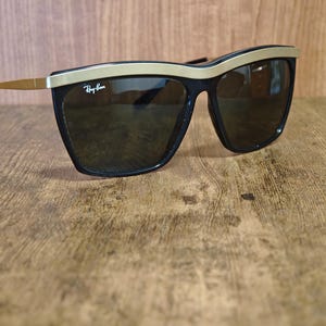 Peut inclure: Lunettes de soleil noires avec une monture sup&eacute;rieure et des branches dor&eacute;es. Les lunettes ont des verres fonc&eacute;s et le logo Ray-Ban est visible sur le verre sup&eacute;rieur gauche. Les lunettes sont pos&eacute;es sur un fond en bois.