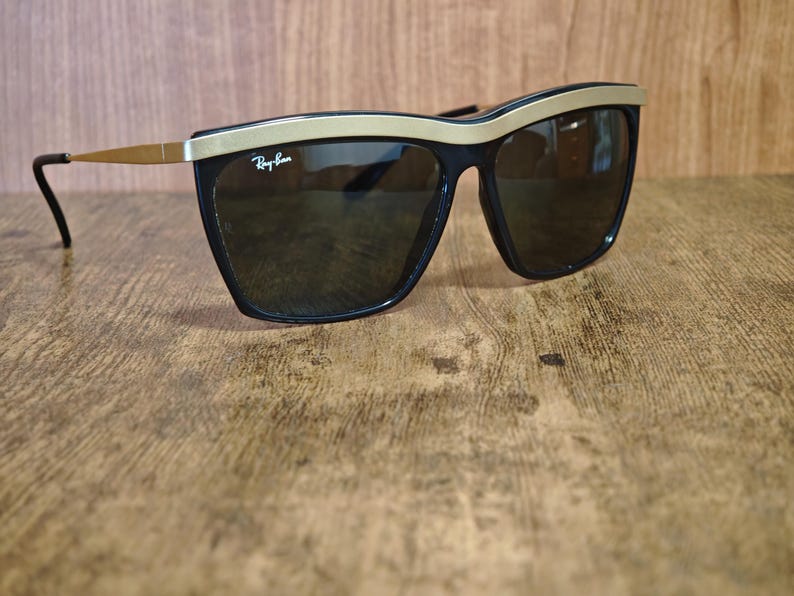 Peut inclure: Lunettes de soleil noires avec une monture sup&eacute;rieure et des branches dor&eacute;es. Les lunettes ont des verres fonc&eacute;s et le logo Ray-Ban est visible sur le verre sup&eacute;rieur gauche. Les lunettes sont pos&eacute;es sur un fond en bois.