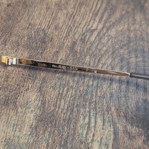May include: Close-up of a gold-toned eyeglass arm with the text "B&L RAY-BAN" etched into the metal. The arm has a dark brown tip and is set against a textured, brown wooden surface.
