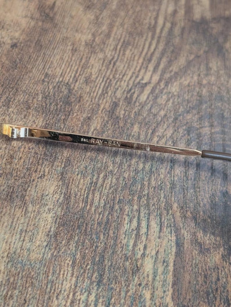 May include: Close-up of a gold-toned eyeglass arm with the text "B&L RAY-BAN" etched into the metal. The arm has a dark brown tip and is set against a textured, brown wooden surface.