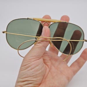 May include: Vintage aviator sunglasses with gold-tone frames and green lenses. The sunglasses feature a double bridge and a light-colored brow bar. The lenses are oval-shaped and the arms curve around the ears.