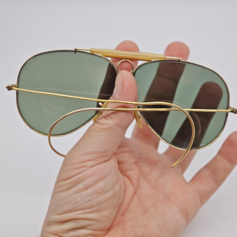 May include: Vintage aviator sunglasses with gold-tone frames and green lenses. The sunglasses feature a double bridge and a light-colored brow bar. The lenses are oval-shaped and the arms curve around the ears.
