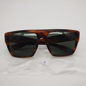 May include: Brown tortoiseshell sunglasses with dark lenses. The sunglasses have a rectangular shape with a thick frame. The lenses are dark green. The sunglasses are on a white surface.