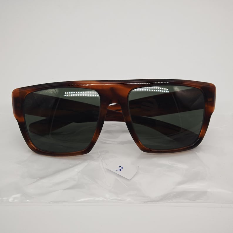 May include: Brown tortoiseshell sunglasses with dark lenses. The sunglasses have a rectangular shape with a thick frame. The lenses are dark green. The sunglasses are on a white surface.