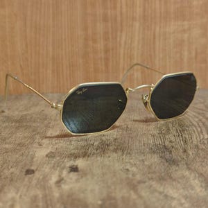 May include: Gold-framed sunglasses with dark lenses. The sunglasses have an octagonal shape and the Ray-Ban logo is visible on the lens. The frame is a thin, delicate gold color.