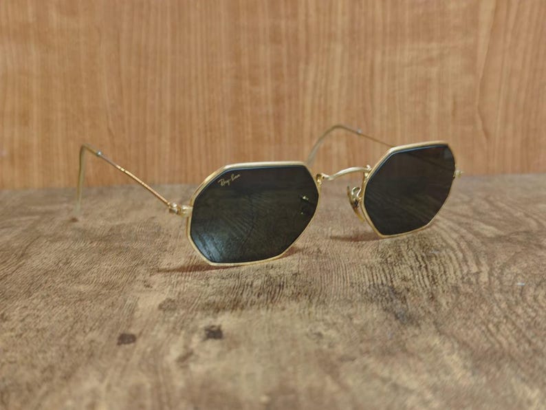 May include: Gold-framed sunglasses with dark lenses. The sunglasses have an octagonal shape and the Ray-Ban logo is visible on the lens. The frame is a thin, delicate gold color.