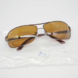 May include: Brown aviator sunglasses with a double bridge and brown tinted lenses. The sunglasses have a metal frame and the Ray-Ban logo is visible on the left lens. The number "807" is written on the plastic bag.